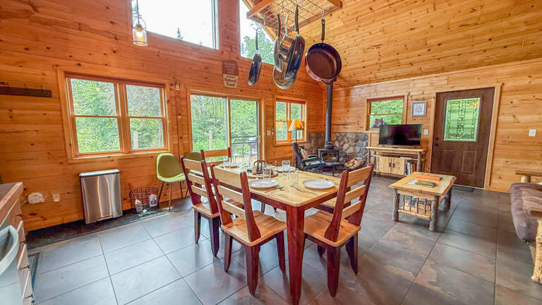 Gather around the handcrafted dining table in this sun-drenched open-concept space, where floor-to-ceiling windows frame forest views. The cozy stone fireplace and rustic wood walls create your perfect mountain retreat atmosphere.