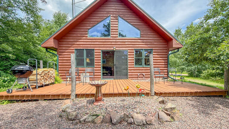 Red log cabin surrounded by lush greenery with spacious deck, BBQ grill, firewood, and outdoor seating, offering a rustic retreat in nature.