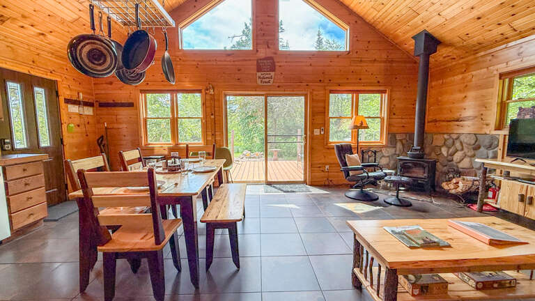 Step into your cozy cabin retreat with soaring vaulted ceilings, where rustic wood beams meet abundant natural light. Gather around the dining table or warm up by the wood-burning stove in this inviting, open-concept space.