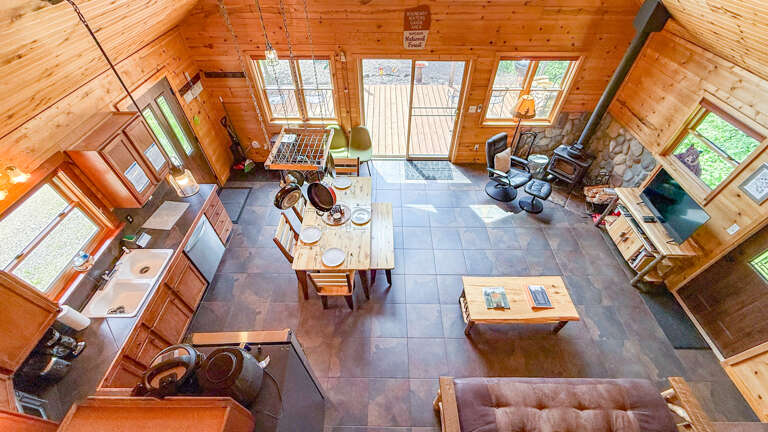 Sunlight streams through vaulted ceilings into your open-concept cabin retreat, where wood beams frame a full kitchen, dining space, and cozy living area with a stone fireplace.