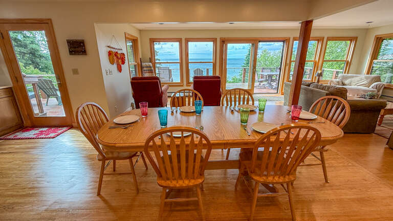 Gather your group around this inviting dining table with colorful glassware, where expansive windows reveal breathtaking water views. The open space flows seamlessly to outdoor decks and cozy seating areas, perfect for memorable meals together.