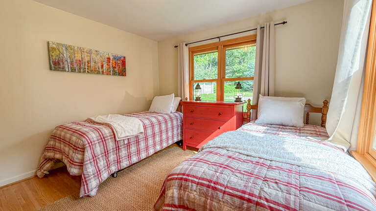 Settle into your peaceful twin bedroom, where cozy plaid bedding and warm natural light through wood-framed windows invite restful sleep surrounded by charming, thoughtful touches.