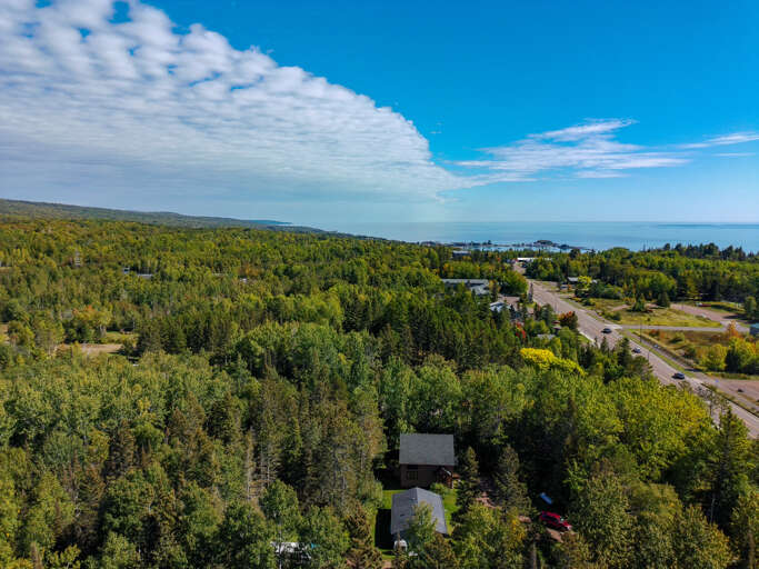 Aerial view showcasing the property nestled within lush forested landscape, with Lake Superior visible on the horizon and a scenic roadway connecting the area.