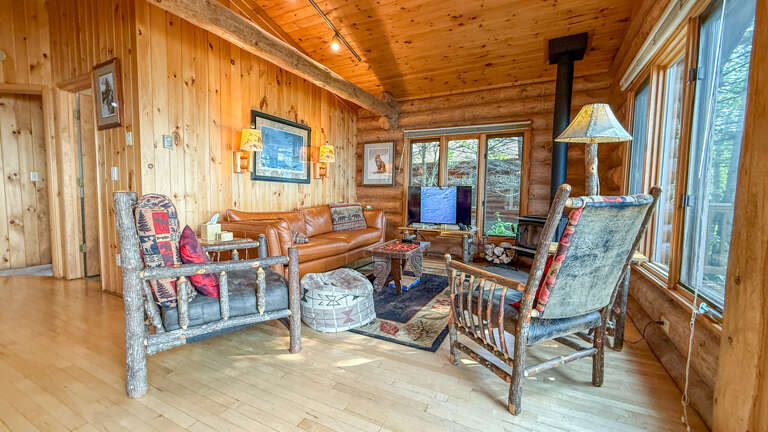 Cozy cabin with rustic log walls, warm leather sofa, and handcrafted chairs around a wood-burning stove. Large windows showcase forest views and flood the space with natural light.