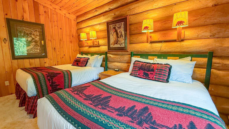 Sink into plush white linens dressed with rustic bear-motif throws, while warm wood walls and gentle lamplight create a cozy cabin retreat perfect for restful mountain nights.