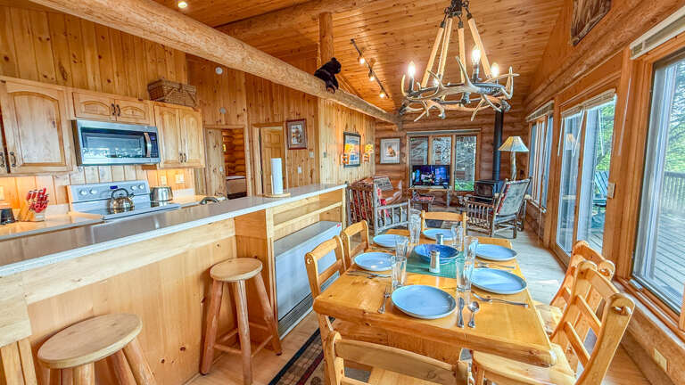 Step into a warm, rustic retreat where the scent of natural wood surrounds you. Your open-concept kitchen and dining area invites cozy meals and laughter under a charming antler chandelier, while windows frame the peaceful forest beyond.