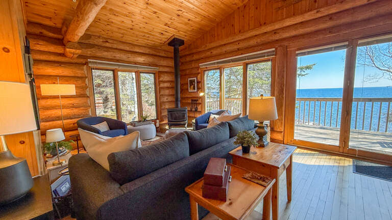 Relax in your cozy log cabin living room with stunning lake views through floor-to-ceiling windows and comfortable seating by the fireplace.