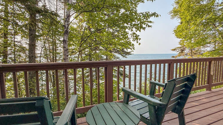 Serene lakeside deck framed by towering trees, offering a peaceful retreat. Green wooden chairs invite you to relax and take in the tranquil water views through the forest.