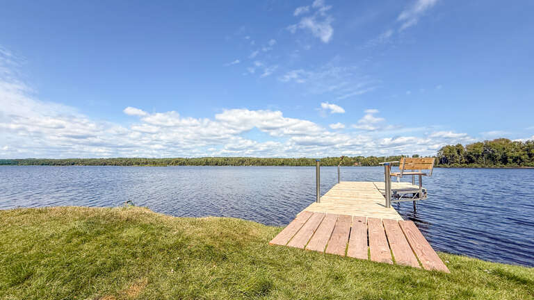 Private dock stretching into tranquil lake waters, with a serene shoreline and lush greenery surrounding the property under open skies.