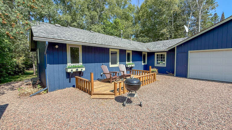 Charming blue-sided retreat with welcoming front deck surrounded by lush trees and gravel driveway. The outdoor seating and BBQ set the stage for relaxing gatherings.