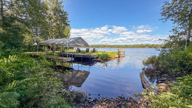 A peaceful lakefront setting, complete with private dock and shaded waterside seating overlooking calm, reflective waters surrounded by lush forest.