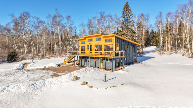 Modern two-story cabin nestled in pristine winter landscape with snow-covered grounds and towering evergreens creating a peaceful retreat setting.