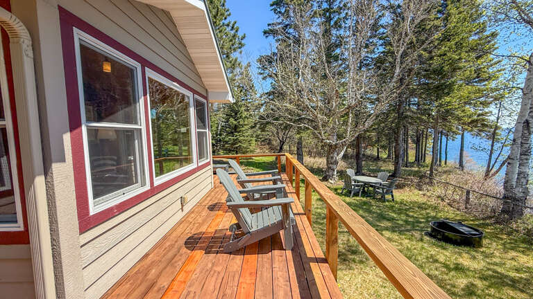 Sunny deck overlooks tranquil lake through tall trees, offering Adirondack chairs for relaxation. Perfect spot to enjoy morning coffee or evening sunsets with serene water views.