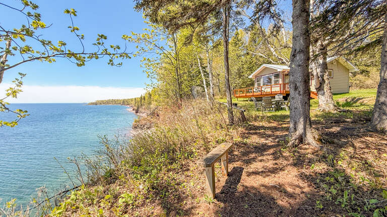 Waterfront cabin with scenic views of Lake Superior, surrounded by tall pines and natural landscape. Path leads down to the lakeshore through wooded property. Spring season visible with emerging foliage.