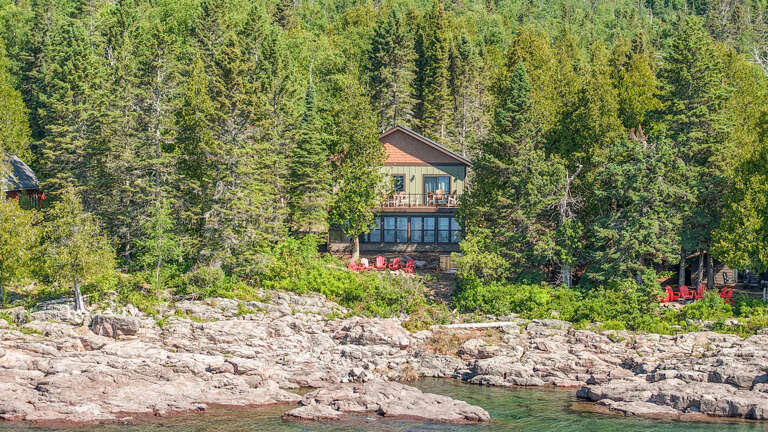 This charming waterfront property is nestled in lush forest with direct rocky shoreline access, offering a peaceful natural escape surrounded by evergreens and vibrant seasonal foliage.