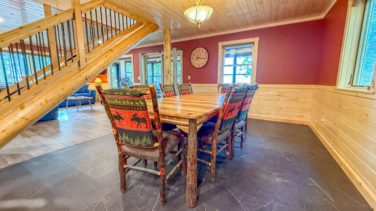 Gather around this rustic wood dining table with colorful patterned chairs in a warm, light-filled cabin setting. The vaulted pine ceiling and cozy red accent wall make every meal feel like a retreat.