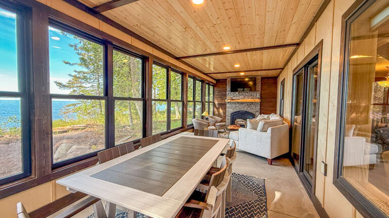 Your lakeside sunroom beckons with panoramic water views through floor-to-ceiling windows, a cozy stone fireplace, and a welcoming dining table perfect for memorable meals with loved ones.