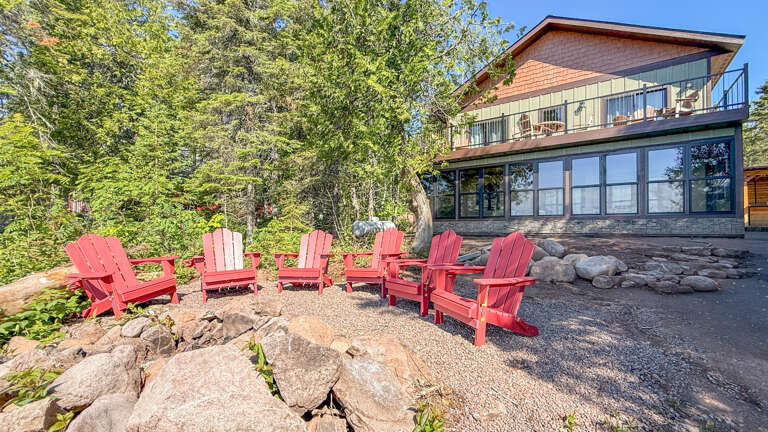 Modern lakefront retreat featuring a spacious upper deck and a gravel patio with six vibrant red Adirondack chairs, nestled among mature trees and natural stone formations in a tranquil waterfront setting.