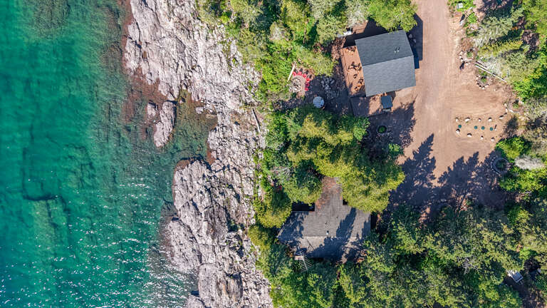 Aerial view of a waterfront vacation rental nestled among trees on a stunning rocky shoreline, with crystal-clear turquoise water and private access to the beach.