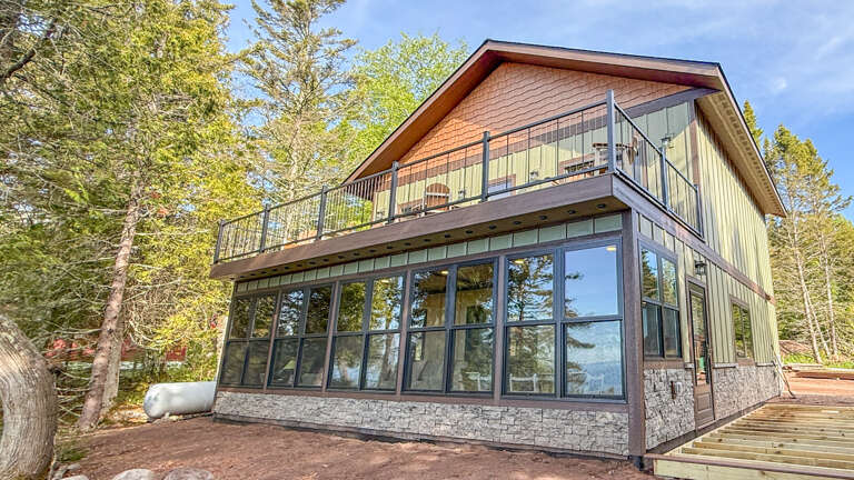 A modern two-story vacation home nestled among trees, featuring expansive windows and an upper balcony, surrounded by wooded landscape and natural light.