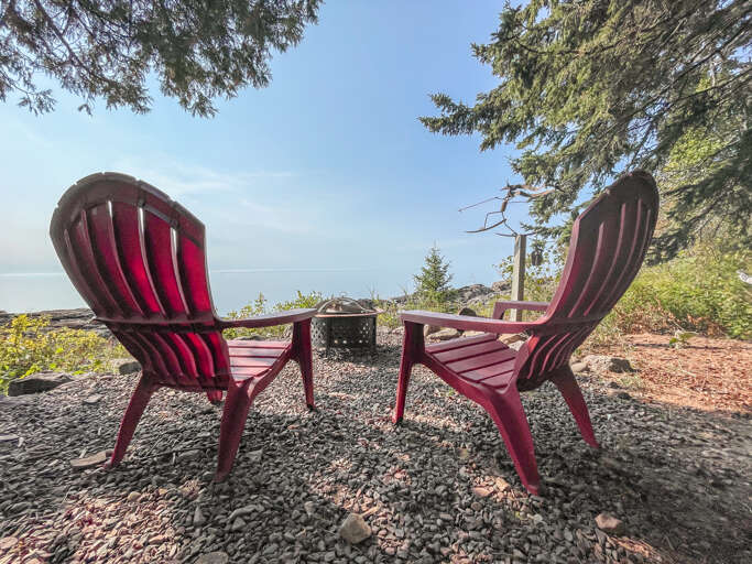 Settle into red Adirondack chairs framing a fire pit, where sea views and towering pines create your perfect retreat for morning coffee or starlit conversations.