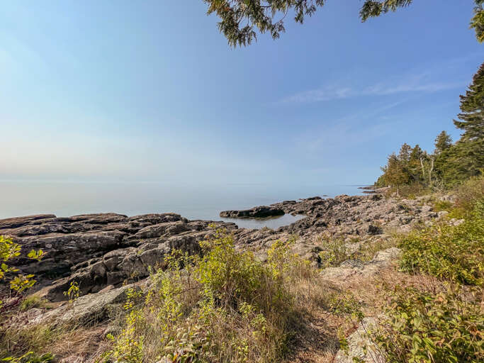 Rocky shoreline framed by towering evergreens, with calm waters stretching into the horizon—a serene natural backdrop near the property.