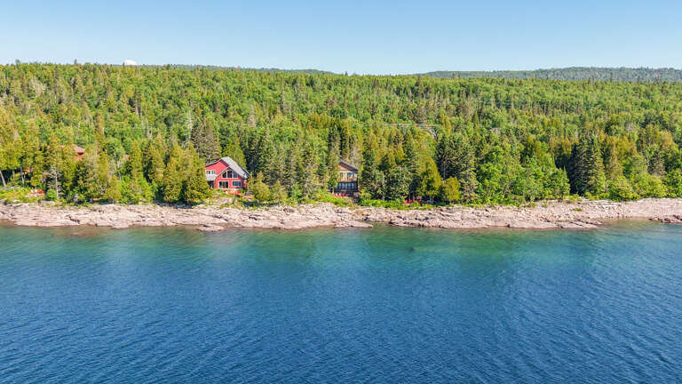 Aerial view of secluded waterfront cabins nestled among dense evergreen forest along a rocky shoreline, offering a peaceful natural retreat in a pristine wooded setting.