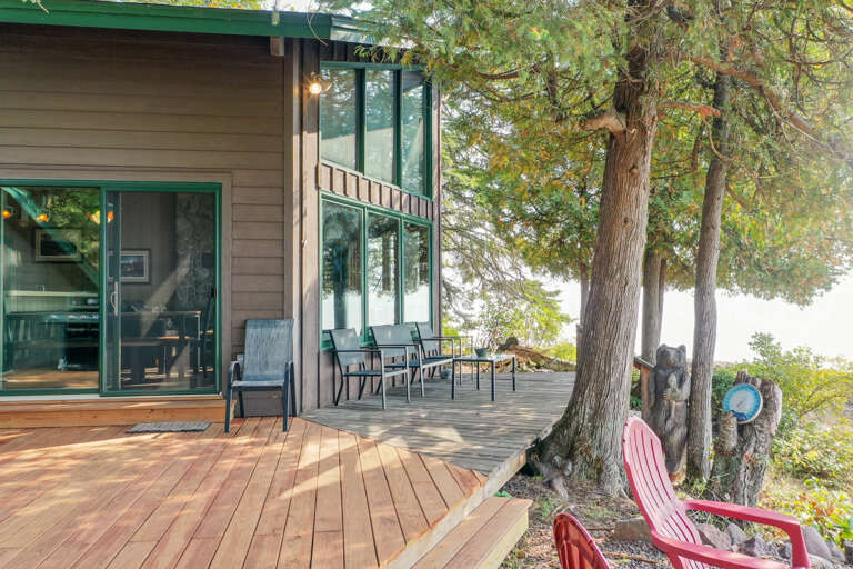 A spacious wooden deck with comfortable seating nestled among mature trees, offering waterfront views and seamless access to the modern interior through large glass doors.
