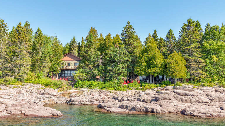A waterfront property nestled among towering pines and vibrant greenery, set on a dramatic rocky shoreline with crystal-clear waters nearby.