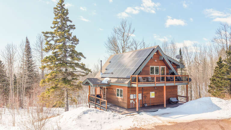 A cozy winter cabin nestled among snow-covered evergreens, featuring solar panels and multiple decks for mountain relaxation.