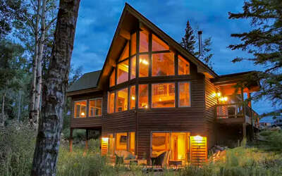 Modern lakeside cabin with floor-to-ceiling windows glowing warmly against the evening sky, surrounded by towering birch and pine trees.