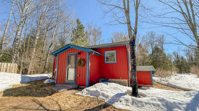 A charming red cabin nestled in snow-covered woods, offering a cozy winter retreat surrounded by bare trees and evergreens under clear blue skies.