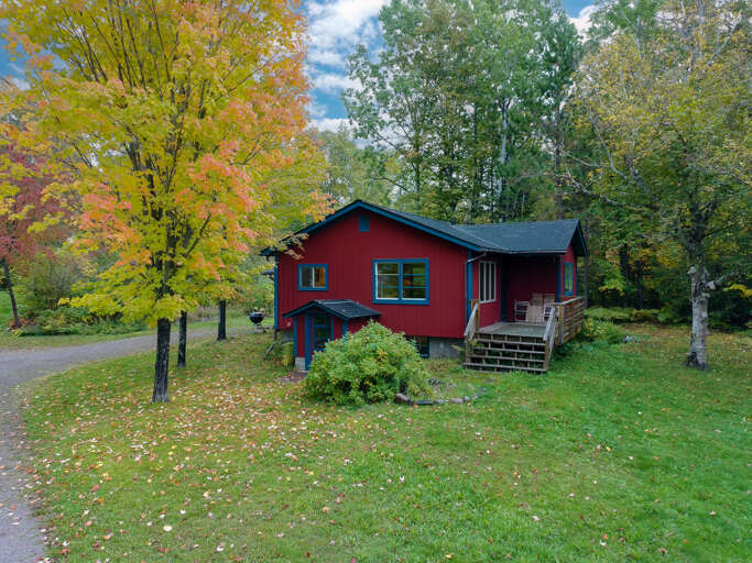 This charming red cottage sits nestled among mature trees with vibrant fall foliage, offering a peaceful autumn retreat on spacious grounds.