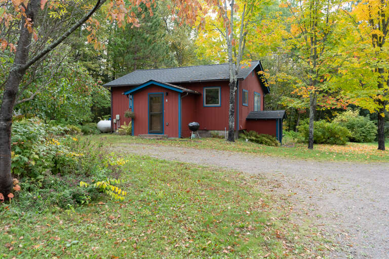 A charming red cabin nestled among vibrant autumn foliage, with a gravel drive and grill ready for your outdoor gatherings in a peaceful wooded setting.