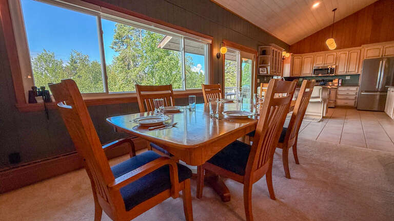 Gather around this warm dining table where sunlight streams through floor-to-ceiling windows, revealing lush treetops and clear skies. The open kitchen layout invites you to cook and connect with ease.