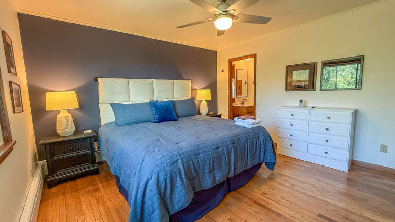 Unwind in this serene bedroom featuring a plush bed with calming blue linens, warm bedside lighting, and a ceiling fan to keep you comfortable. Your private ensuite awaits through the doorway.
