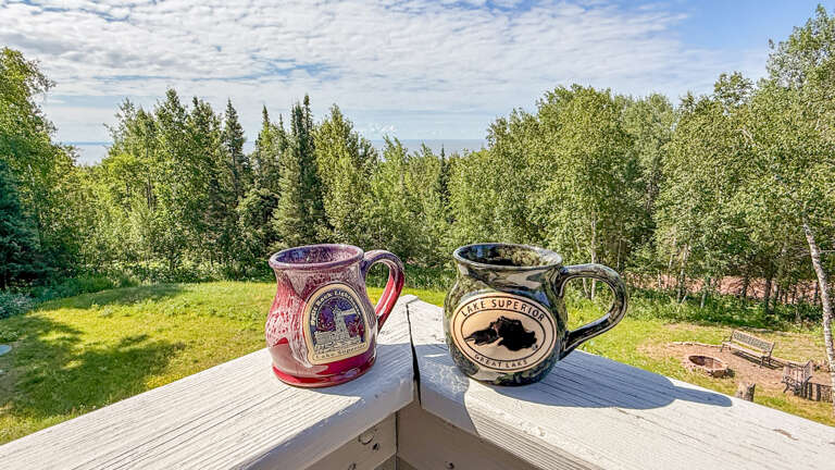 Two handcrafted mugs rest on a weathered deck railing, framed by lush forest and open sky—a serene spot to sip your morning coffee and soak in the quiet beauty around you.