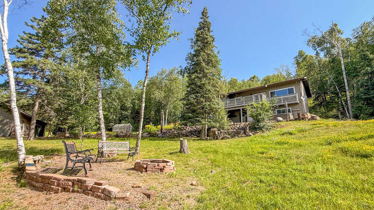 A peaceful woodland retreat with private fire pit and seating area, surrounded by towering birch and evergreen trees under bright blue skies.
