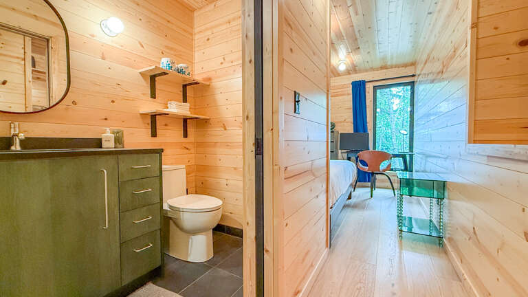 Your cozy cabin features warm wood paneling throughout, with a compact bathroom on the left and a bright bedroom just steps awayânatural light and greenery framed by your window.