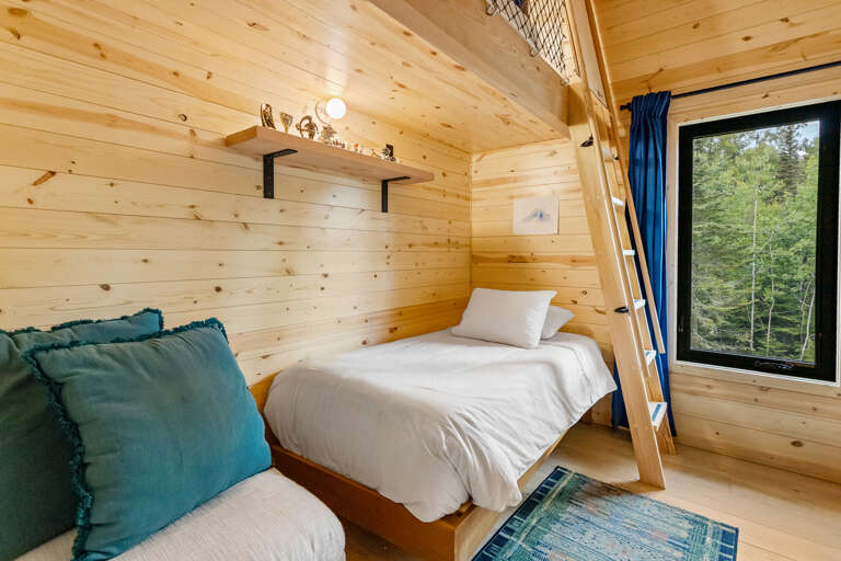 Wake up to crisp forest views from your cozy cabin bedroom, where natural pine walls glow warmly around your peaceful sleeping space. A tucked-away loft adds charm, while sunlight streams through the window framing lush greenery outside.