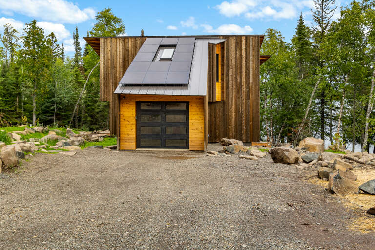 A striking modern property featuring natural wood accents and solar panels, set amid lush forest scenery with a gravel driveway and natural stone landscaping.