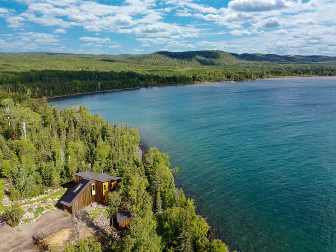 Aerial view of a lakeside cabin nestled among evergreens along pristine turquoise waters, with forested hills and blue skies in the distance.