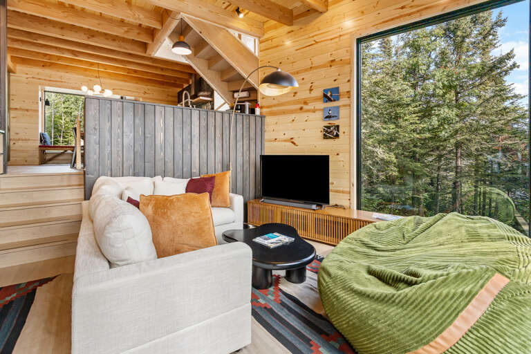 Curl up in your cozy living area with soaring wood beams, plush seating, and a wall of windows framing lush forest views—your perfect retreat.
