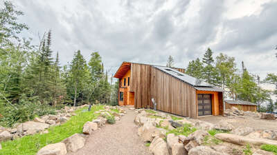 A modern mountain retreat nestled among tall pines and natural stone landscaping, offering a warm welcome to your woodland escape.