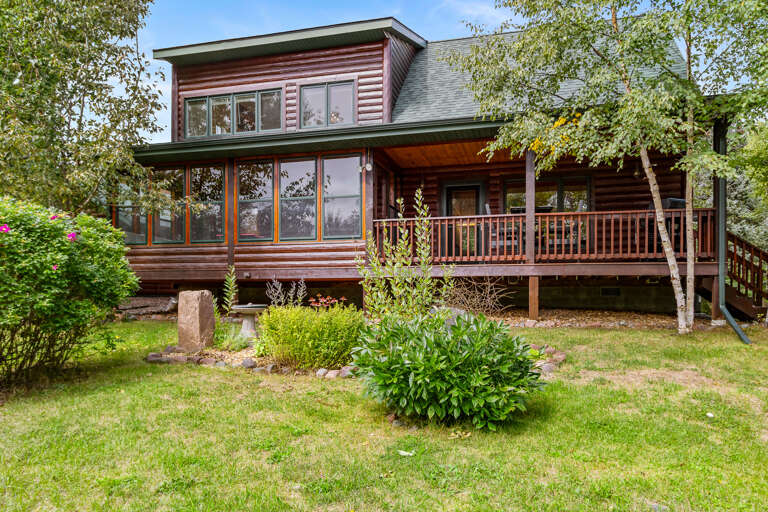 A charming cabin-style retreat with warm wood siding and a spacious wraparound deck, surrounded by lush greenery and mature trees in a peaceful natural setting.