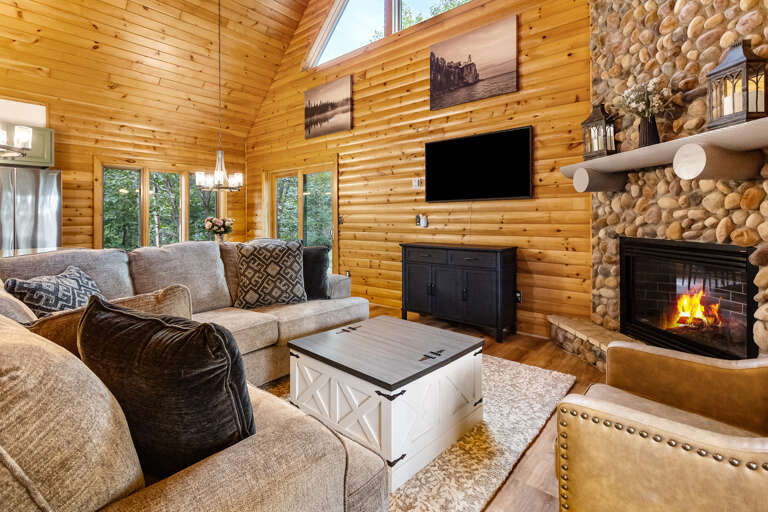 Curl up in your inviting cabin retreat with soaring vaulted ceilings, warm wood paneling, and a stunning stone fireplace crackling beside plush seating, all bathed in natural light from tall windows overlooking the trees.