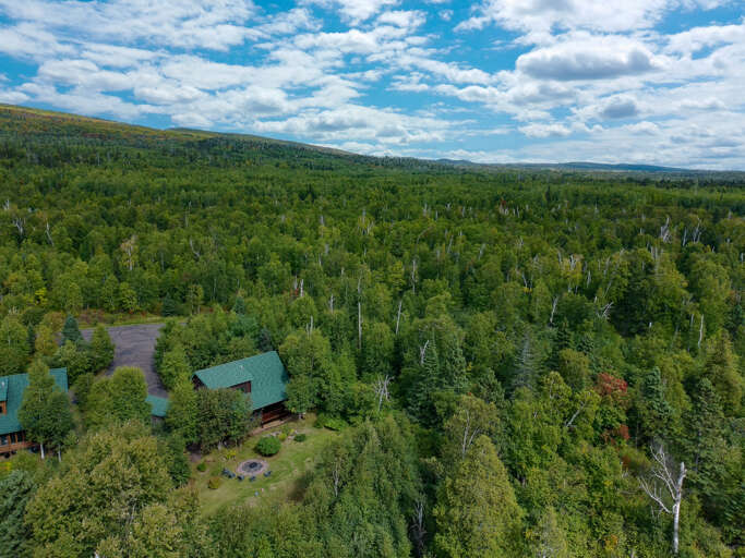 An aerial view reveals the property nestled within vast, pristine forestland beneath expansive skies—a secluded retreat surrounded by nature's tranquility.