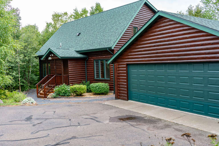 Charming log cabin with green trim and a welcoming covered entry, surrounded by lush forest and natural greenery. Ample parking and garage space await your arrival.