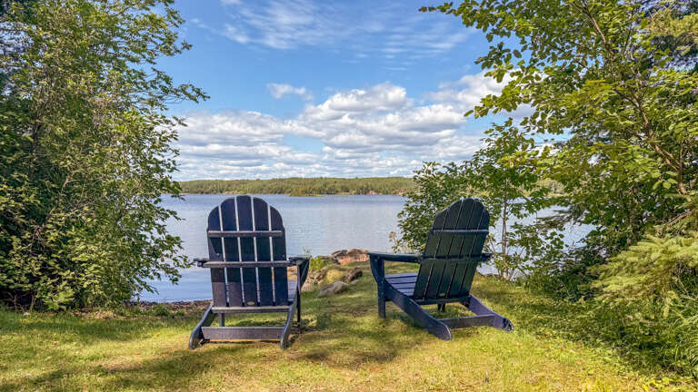 Two Adirondack chairs frame a peaceful lakeside setting, perfect for watching the clouds drift by and soaking in the tranquil water views.