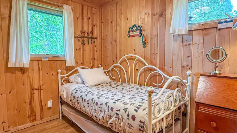 Your charming wood-paneled bedroom features a vintage white metal bed dressed in soft linens, warm natural light from garden-facing windows, and thoughtful touches like a vanity mirror and keepsake dÃ©corâperfectly cozy for restful nights.